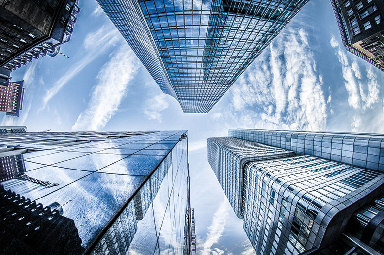 Upward view of modern skyscrapers with reflective glass façades, captured using a wide-angle lens that distorts perspective. The buildings converge dramatically towards a bright, cloud-streaked sky, emphasising height and urban scale.